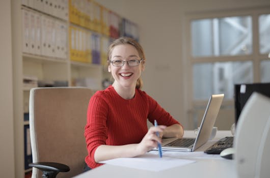 Happy woman in red sweater working at office desk with laptop and pen, smiling at camera.